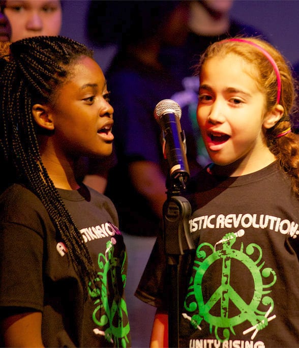 2 children on stage sharing microphone singing together.