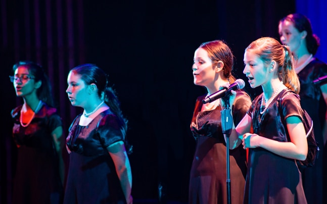 Five girls stand in focus, singing in black performance dresses with a black background and low lighting.
