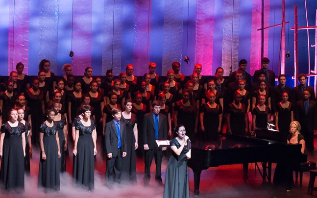 The choir of young singers stand on stage in formation with fog at their feet in front of a blue and pink background. Their is a grand piano, pianist, and a girl with a microphone centerstage.