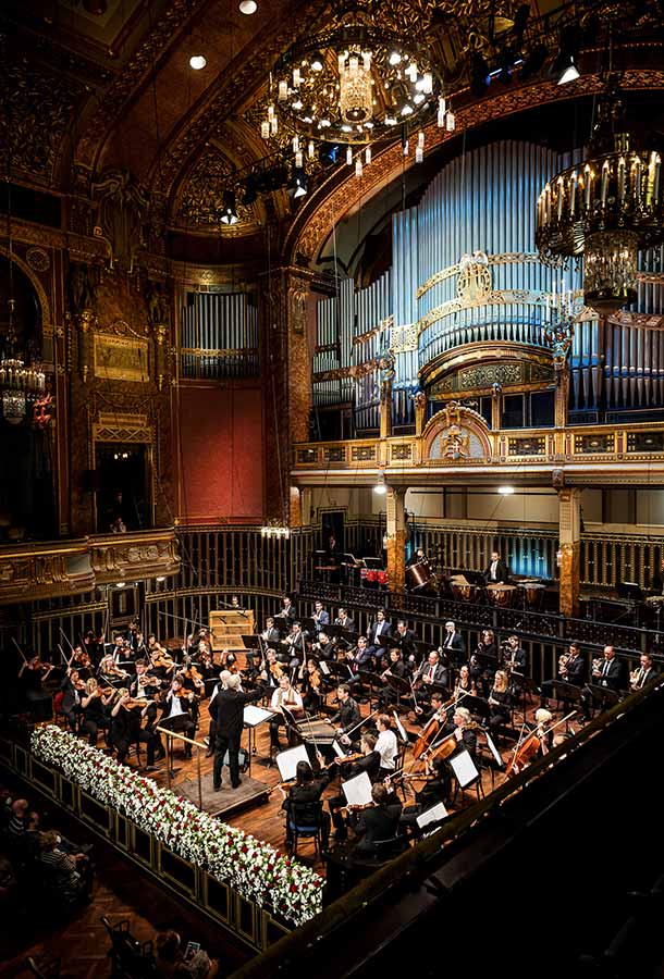 The orchestra is set on a grand stage, performing for an audience as Keller conducts. In the background there is a huge organ and chandeliers.
