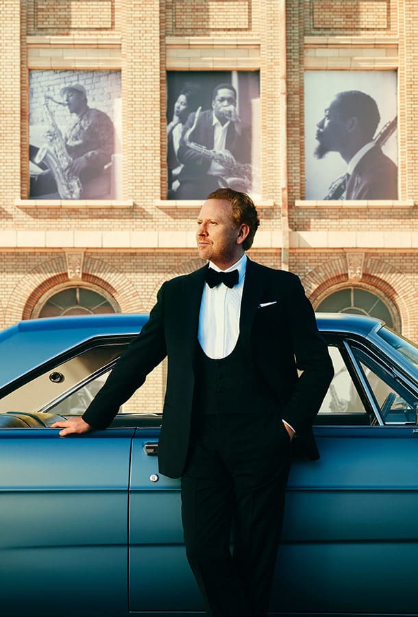 Red-headed man in a black tux leans against an aqua classic car with one hand in his pocket. The backdrop is an old, tan brick building with black and white images of musicians mounted on the outside.