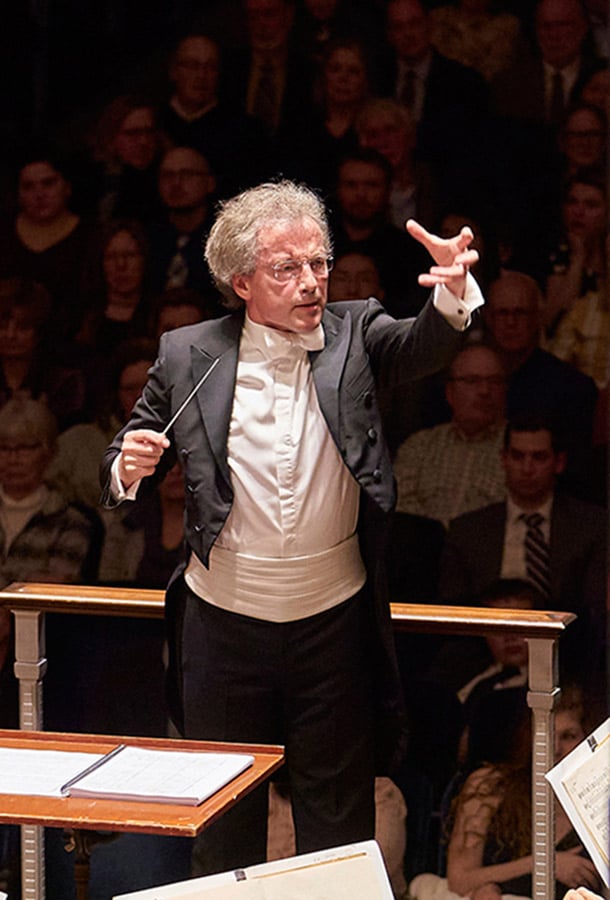 Franz Welser-Möst stands before an audience, conducting passionately with a hand outstretched.