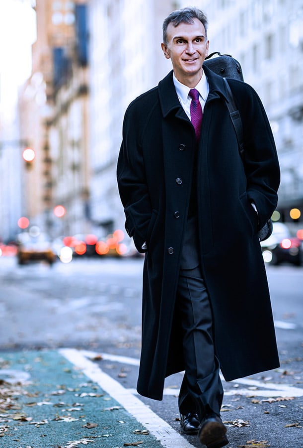 Gil Shaham strolls down a city street wearing a long black coat, a pink tie, and his violin case over his shoulder.