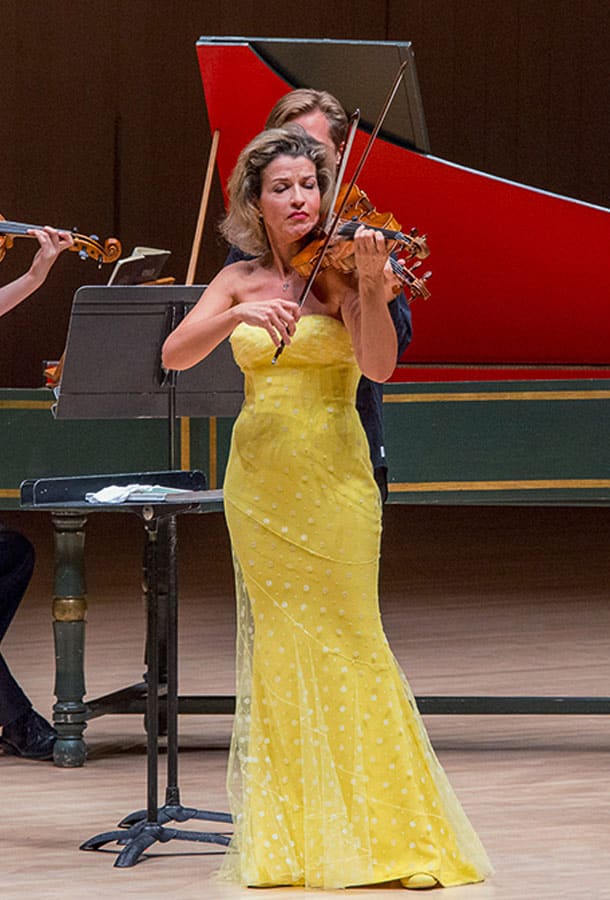 Anne-Sophie Mutter stands, playing her violin. She is center stage, in front of a grand piano, and wearing a floor-length yellow gown.