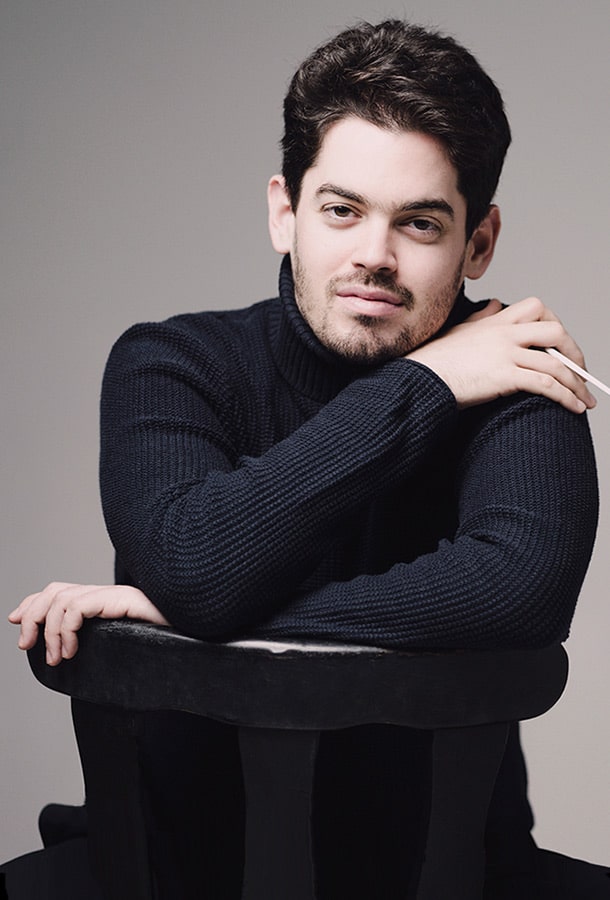 Lahav Shani is pictured, posing in a black turtleneck straddling a chair. He holds his conductor's baton in his right hand, resting on his left shoulder.