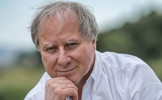 András Keller poses with his chin resting on his hand, looking into the camera. He is wearing a white button-down shirt and is in front of a natural, green background.