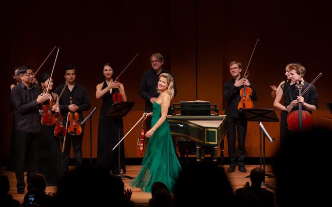 The Mutter Virtuosi stands on stage, each holding their instruments and looking at Anne-Sophie standing in the center wearing a teal dress.
