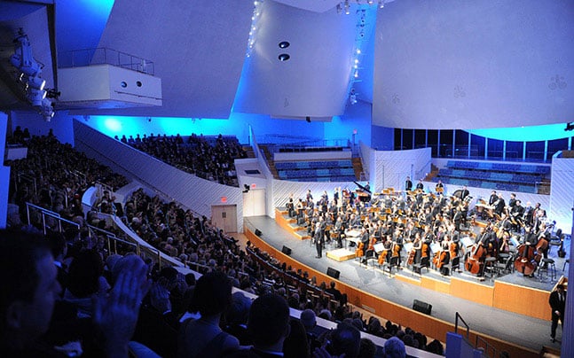 The New World Symphony is on stage illuminated by blue lights and white walls, as a full audience looks on.