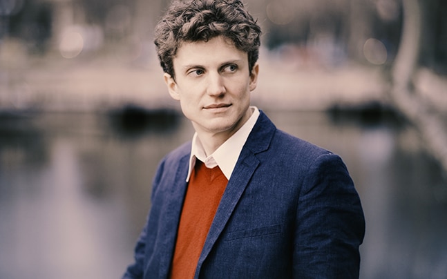Zoltán Fejérvári poses in a navy blazer and burnt-orange sweater, looking off-camera.