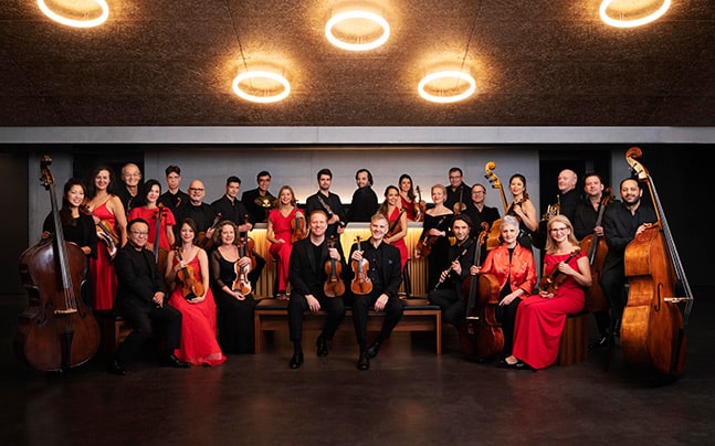 The orchestra poses, smiling with their instruments, all wearing black and red.