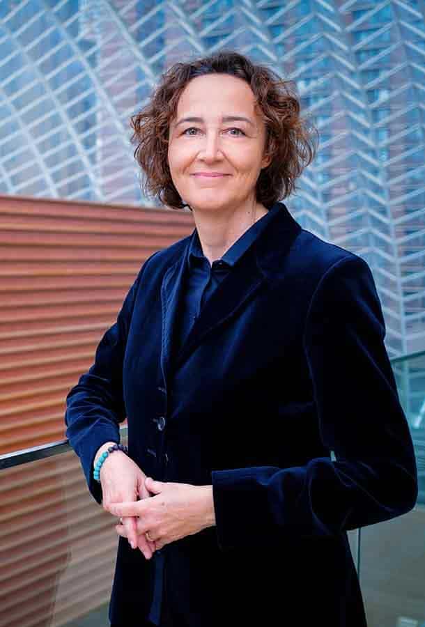 Nathalie Stutzmann smiles into the camera, she is wearing a blue velvet blazer and is in front of a backdrop of blue glass.