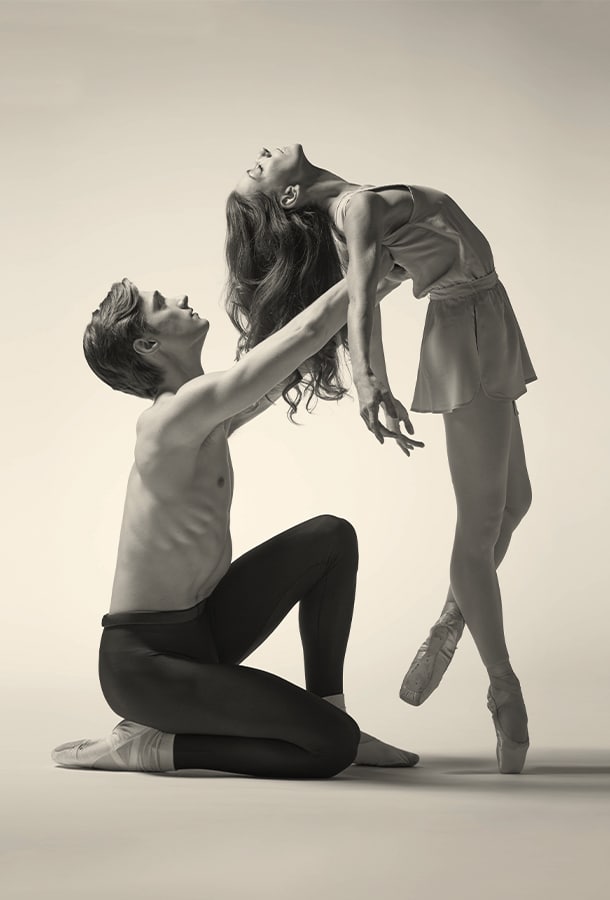 Grey-tone picture of male dancer kneels on the ground, holding up female dancer bending her back into his arms.
