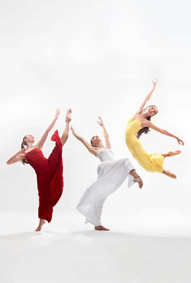 Three woman in flowing dresses of red, white, and yellow pose in various leaps and kicks on a white backdrop.