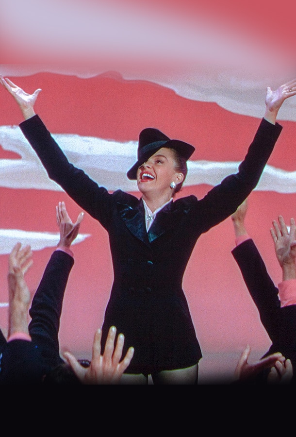 Judy Garland wearing black hat and black dress smiling, posing on stage