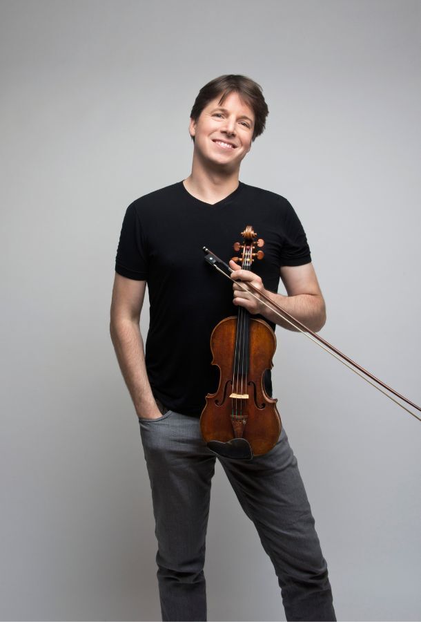 Joshua Bell smiling, holding violin in left hand with his right hand in his pocket.