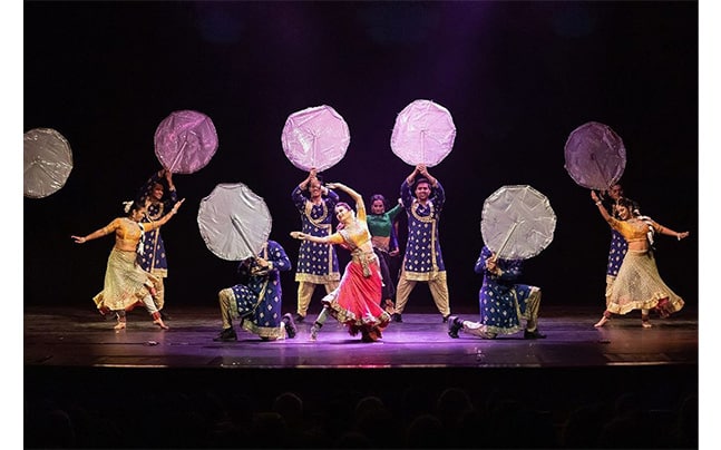 Cast on stage, woman dancing in front surrounded by cast holding drums