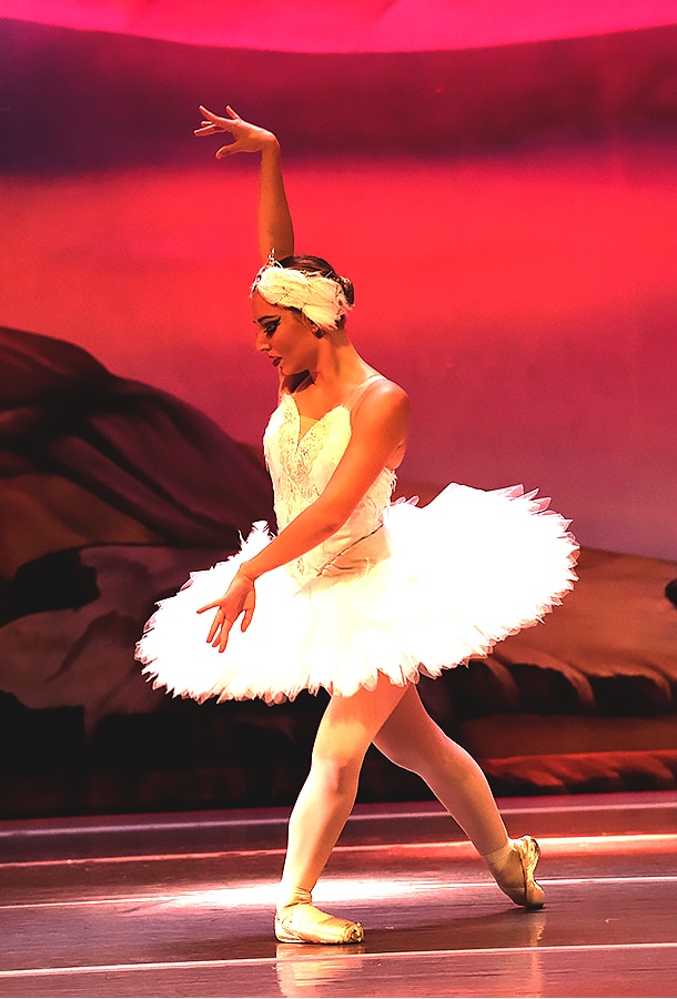 Ballerina in tutu posing on stage with right arm raised.