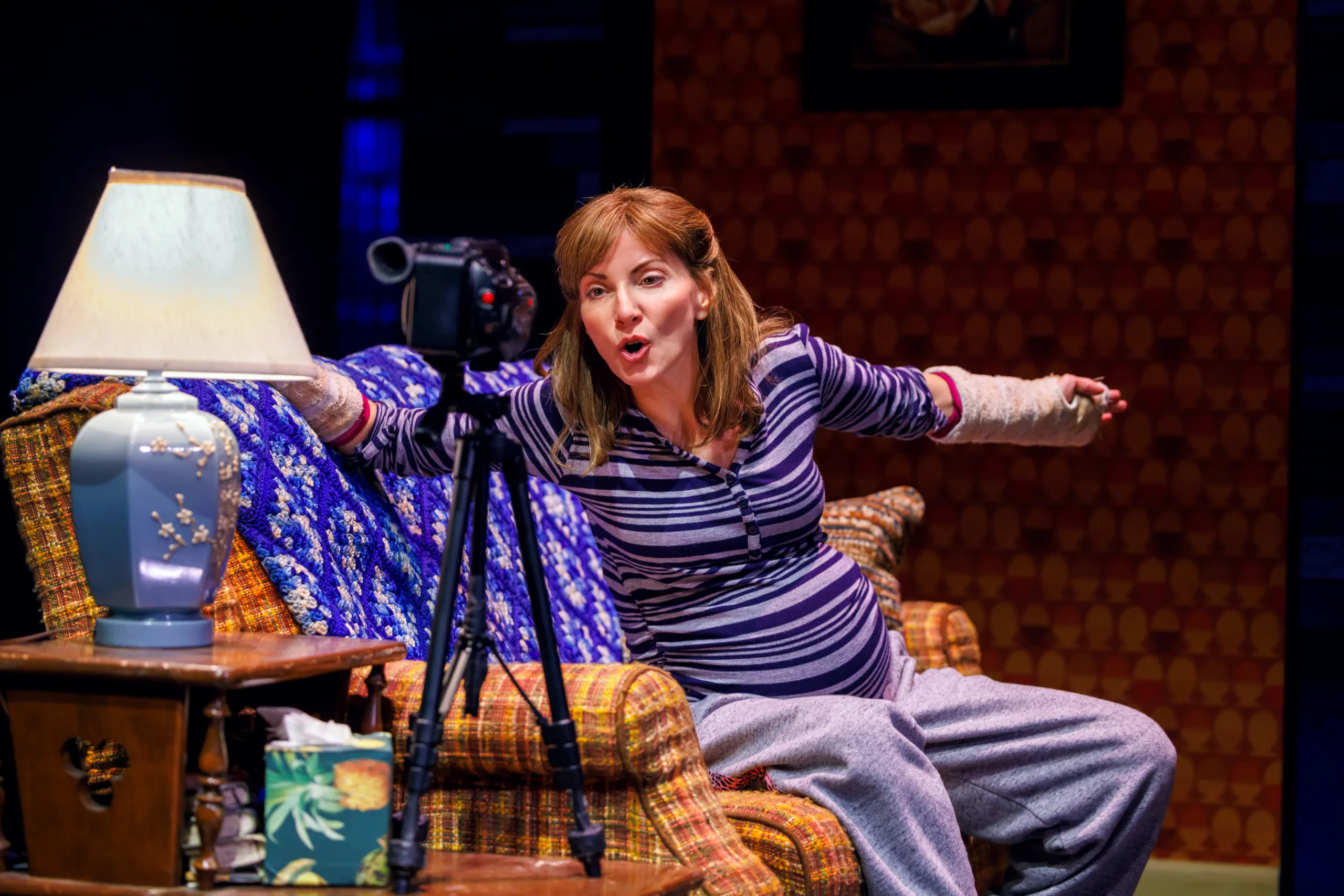 Pattie sitting on the couch in a stripe shirt gesturing toward a camera on a tripod.