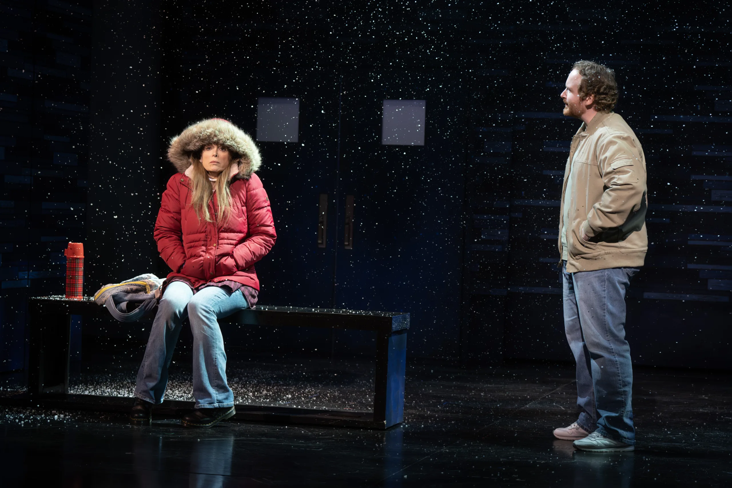 A woman sitting on a bench in a red coat with the hood on and a man standing looking at her.