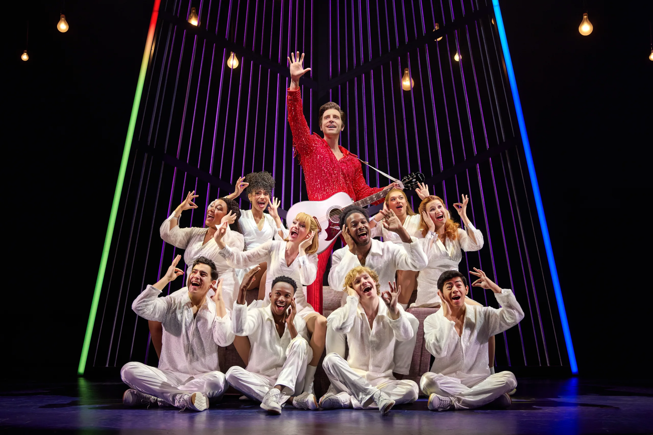 Neil center stage in red with a white acoustic guitar and the company surrounding him dressed in white