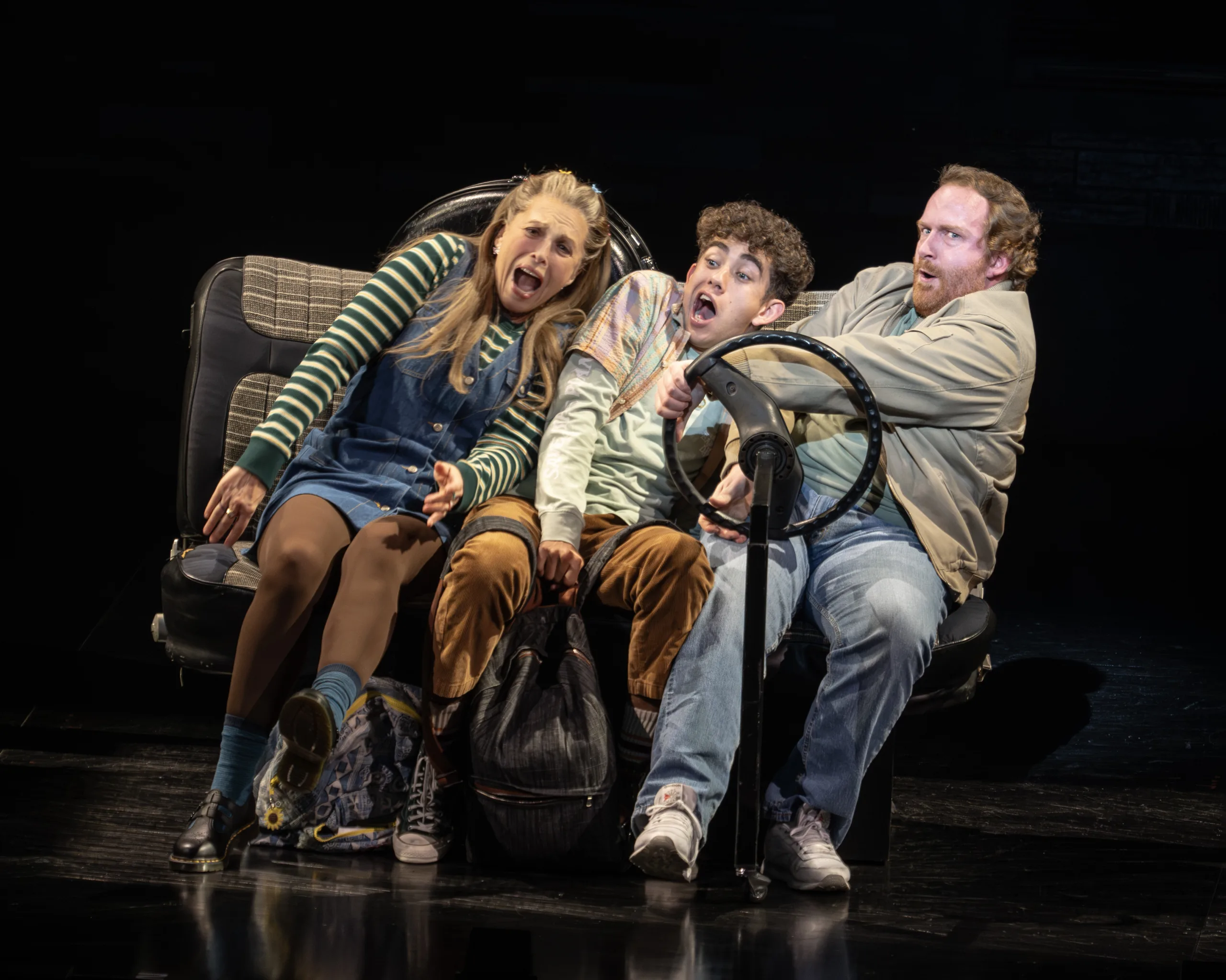 3 people sitting together leaning heavily to the right with an unattached steering wheel in the middle.