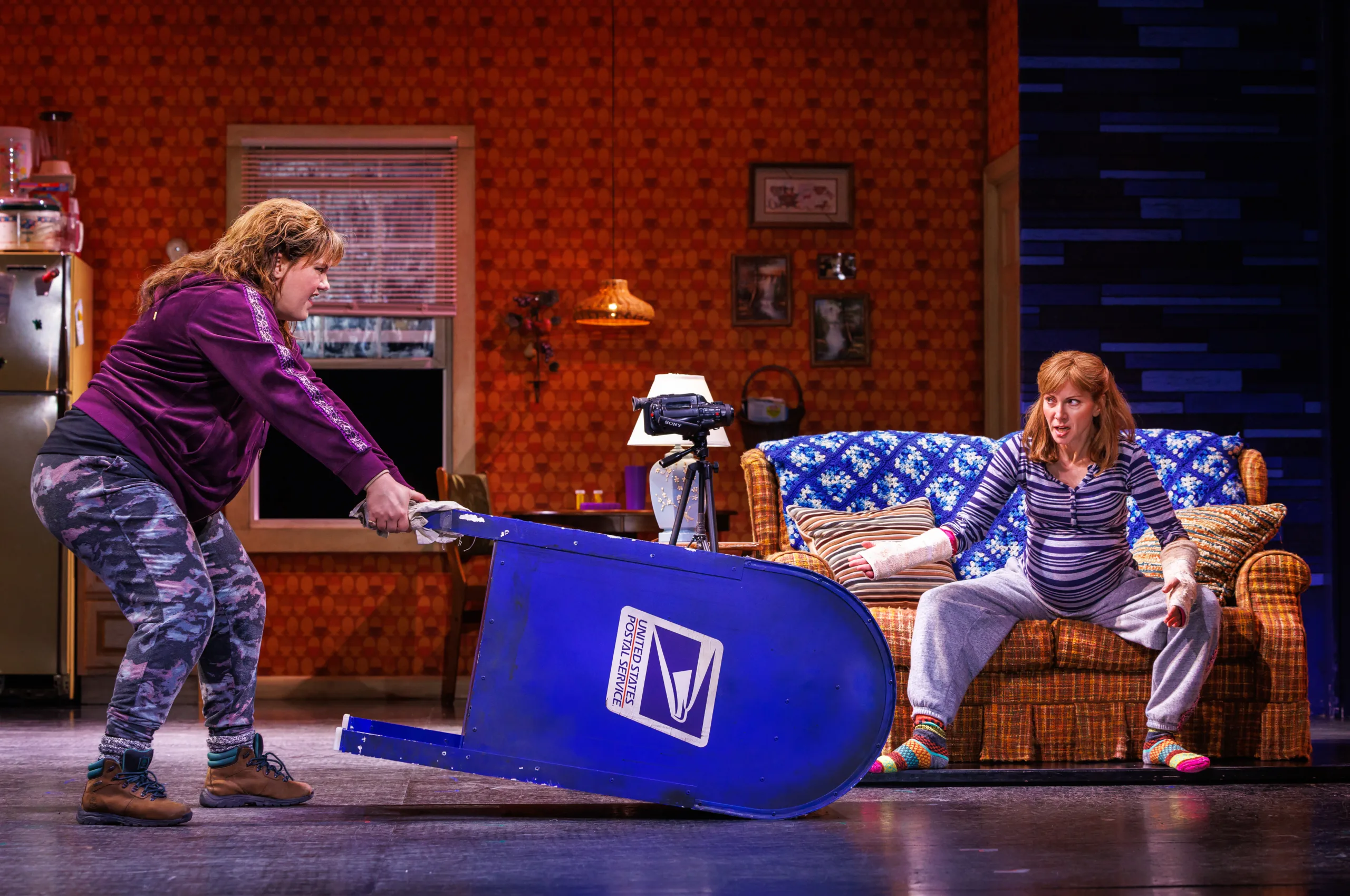 Kimberly dragging a US post mailbox across the stage while pregnant Patties watches from the couch. 