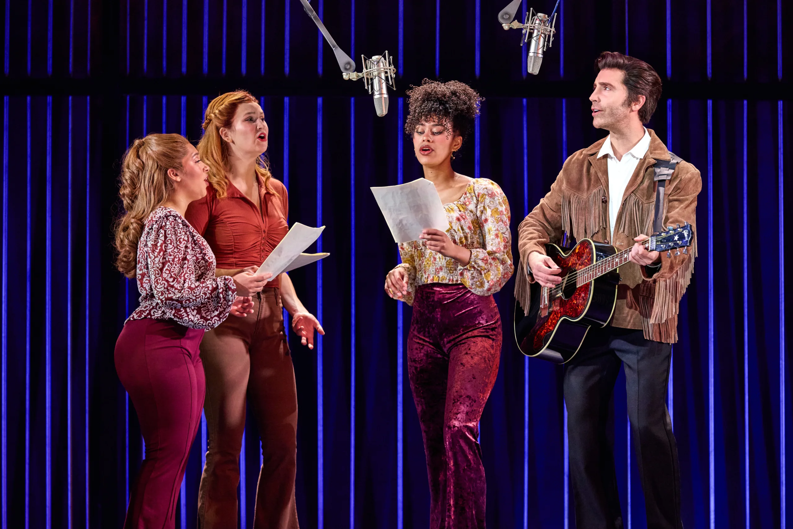 Four people (including Neil playing an acoustic guitar) standing and singing into two hanging microphones.