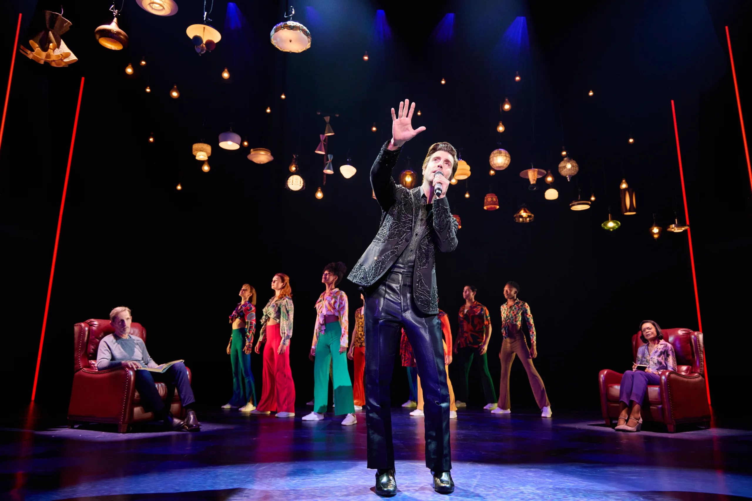 Neil standing in the middle in a black jacket singing into a microphone with his arm outstretched with two people in red armchairs watching him and the company girls in the background. 