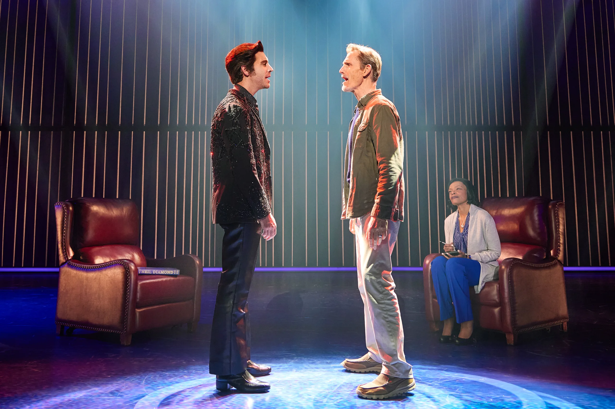 Younger and older Neil Diamond facing each other with a woman in an arm chair watching in the background 