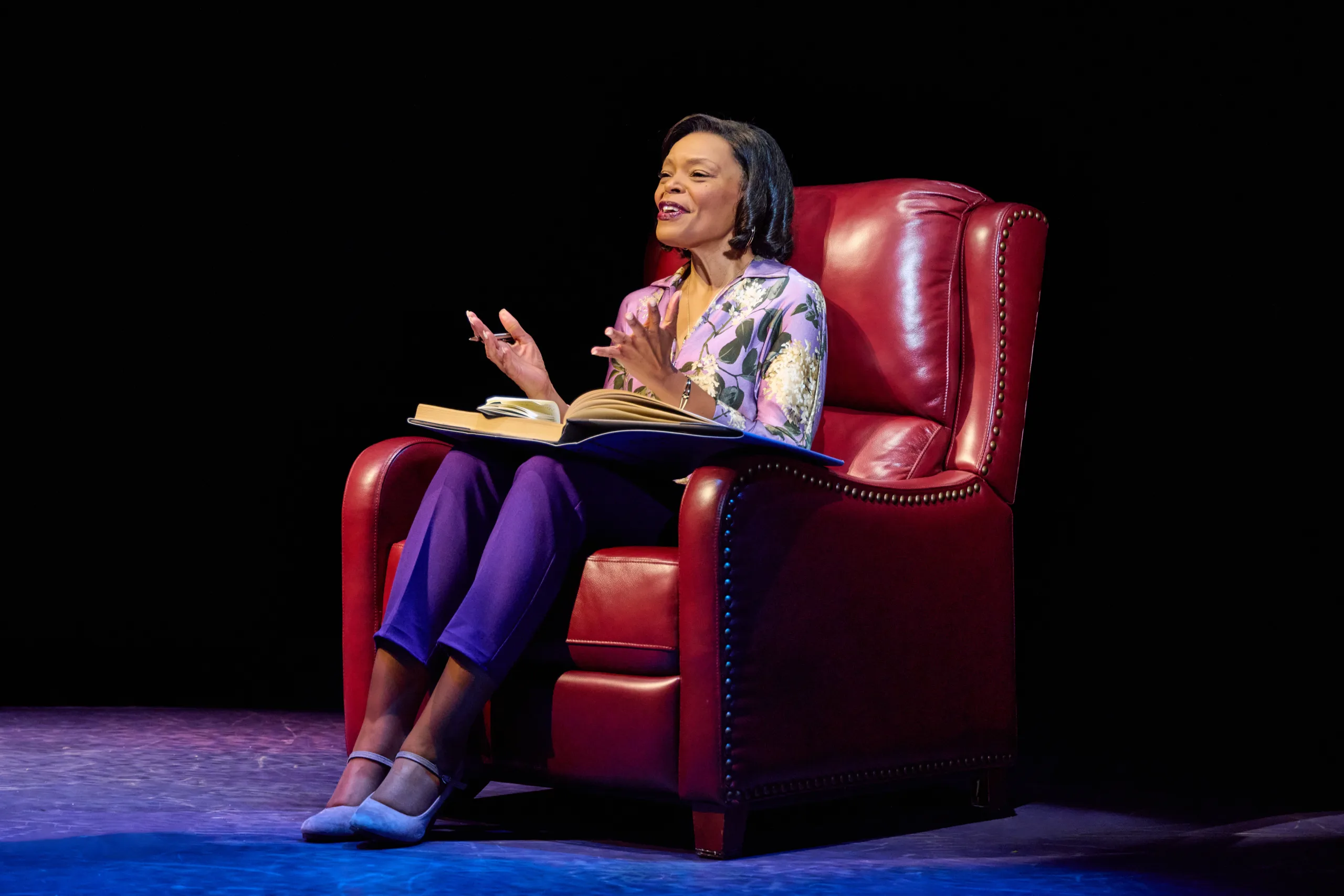 Woman sitting in a red arm chair with a lard book in her lap. 