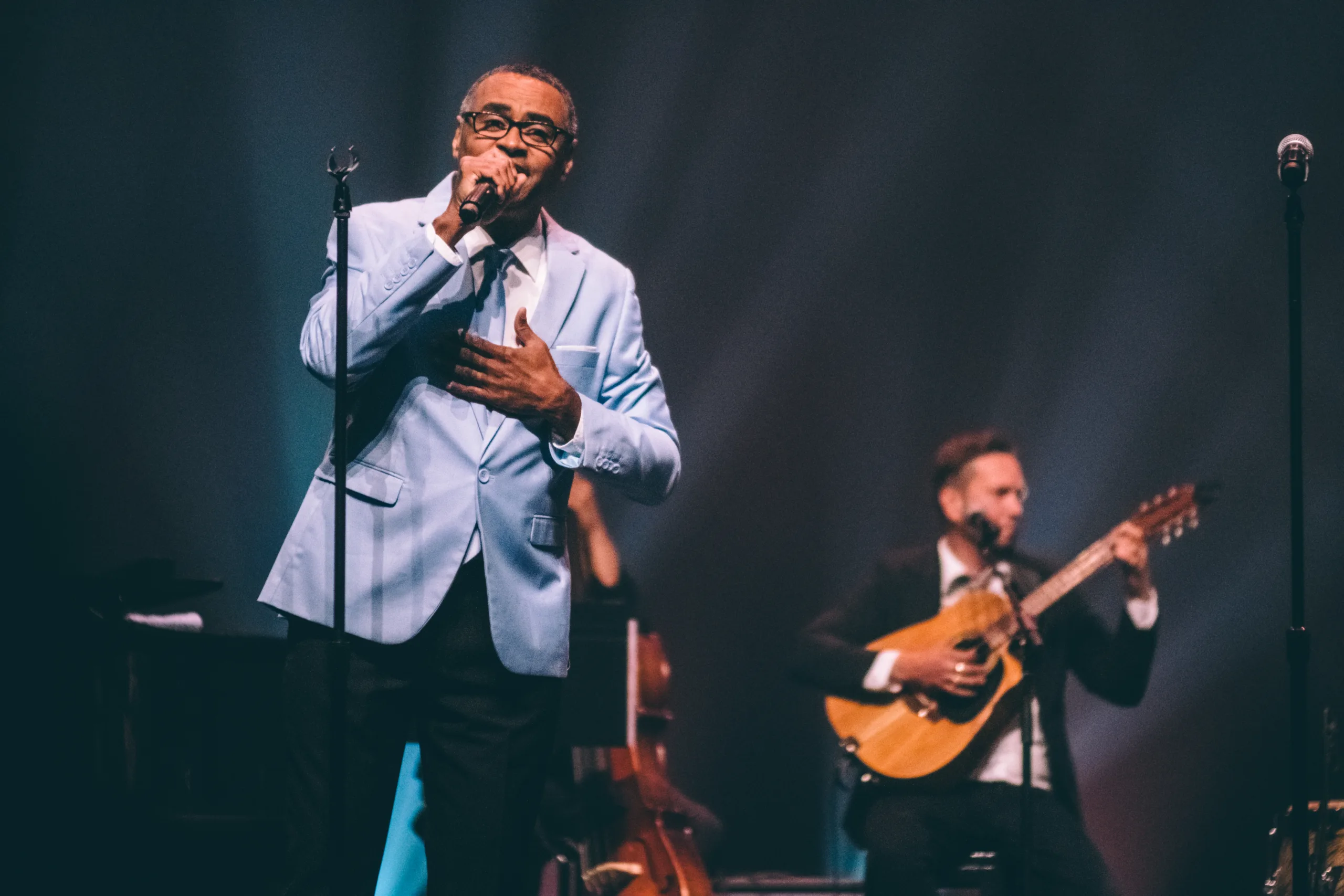 Band member in light blue dinner jacket singing into microphone on stage with guitarist behind him. 