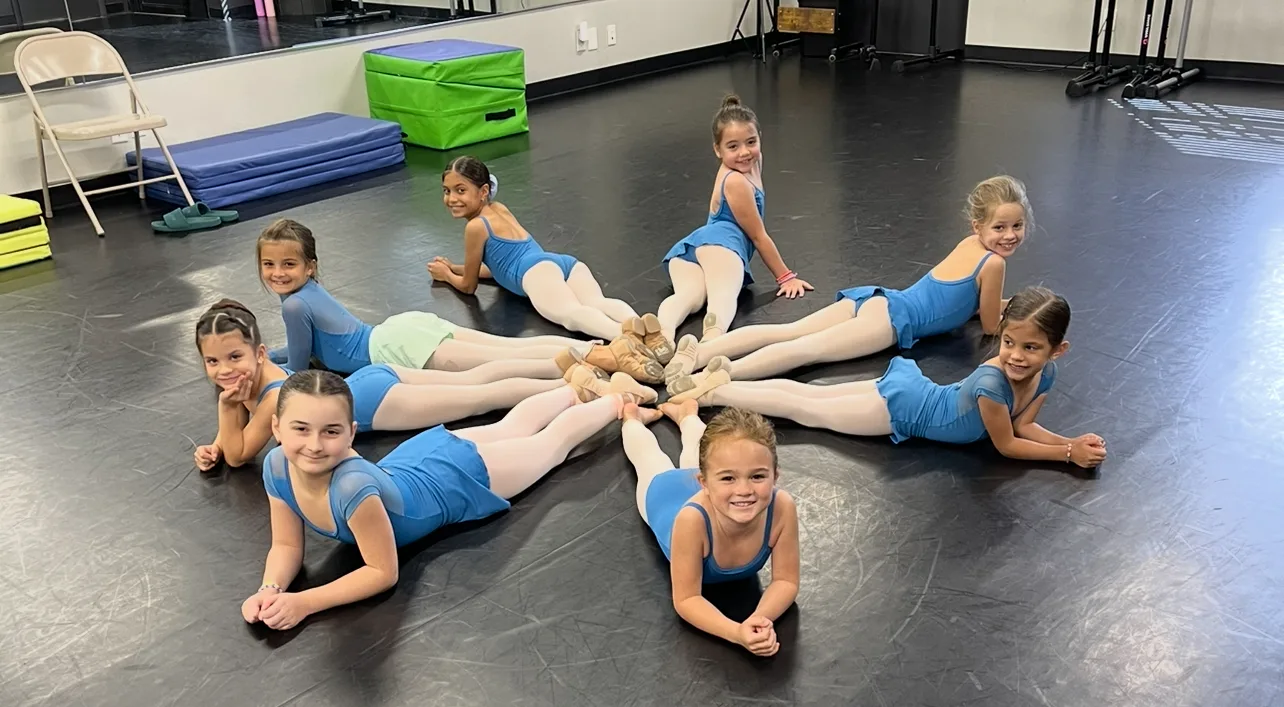 Young girls in blue leotards lay on their stomachs in a circle with toes meeting in the middle. 