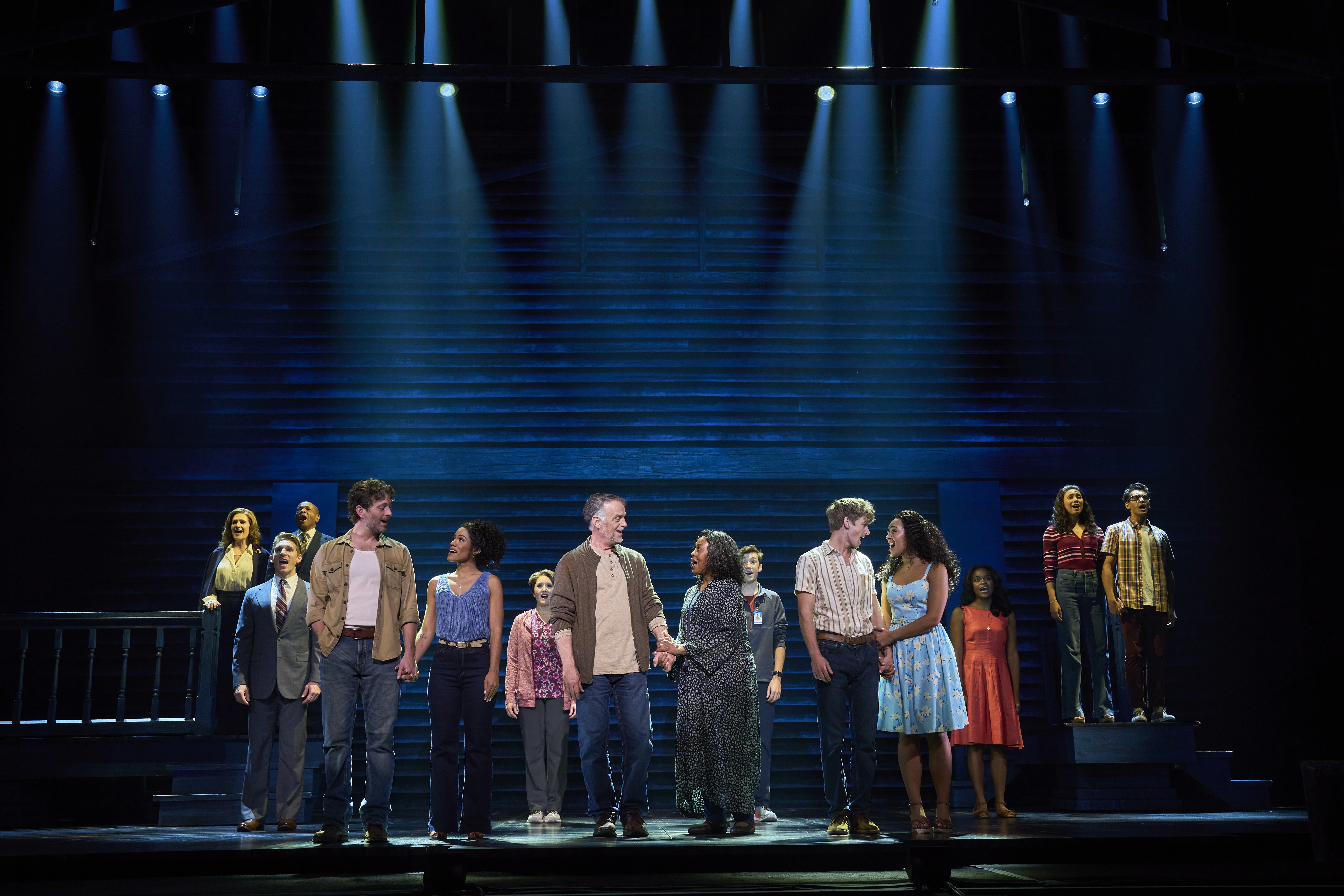 Large ensemble cast standing on stage in a dramatic formation under bright blue spotlights, with actors holding hands in the foreground and others positioned on elevated platforms in the background during a theatrical performance.