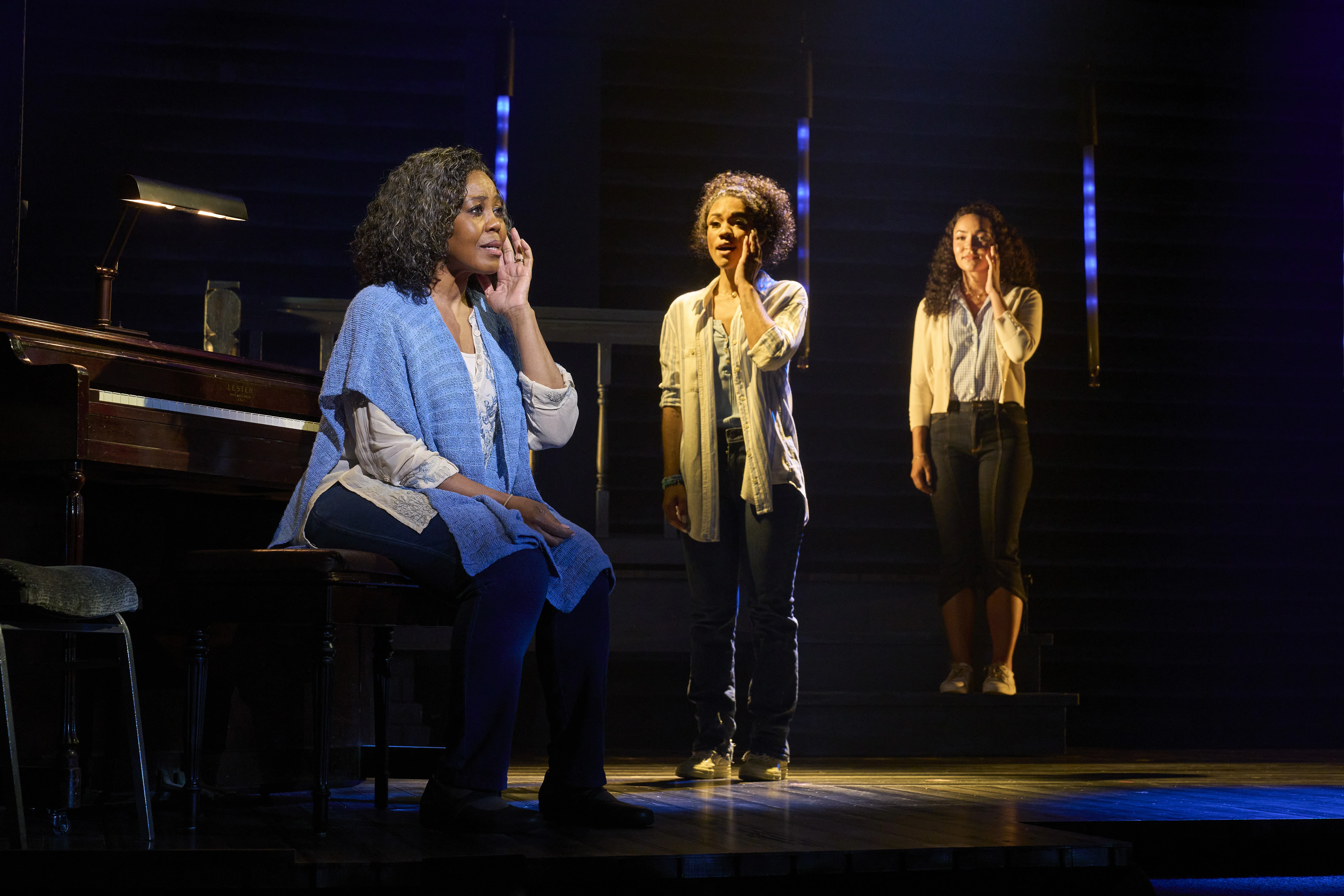 Stage scene from a theatrical production featuring three performers near a piano under dramatic lighting, with two standing in the background and one seated in the foreground wearing a blue shawl.