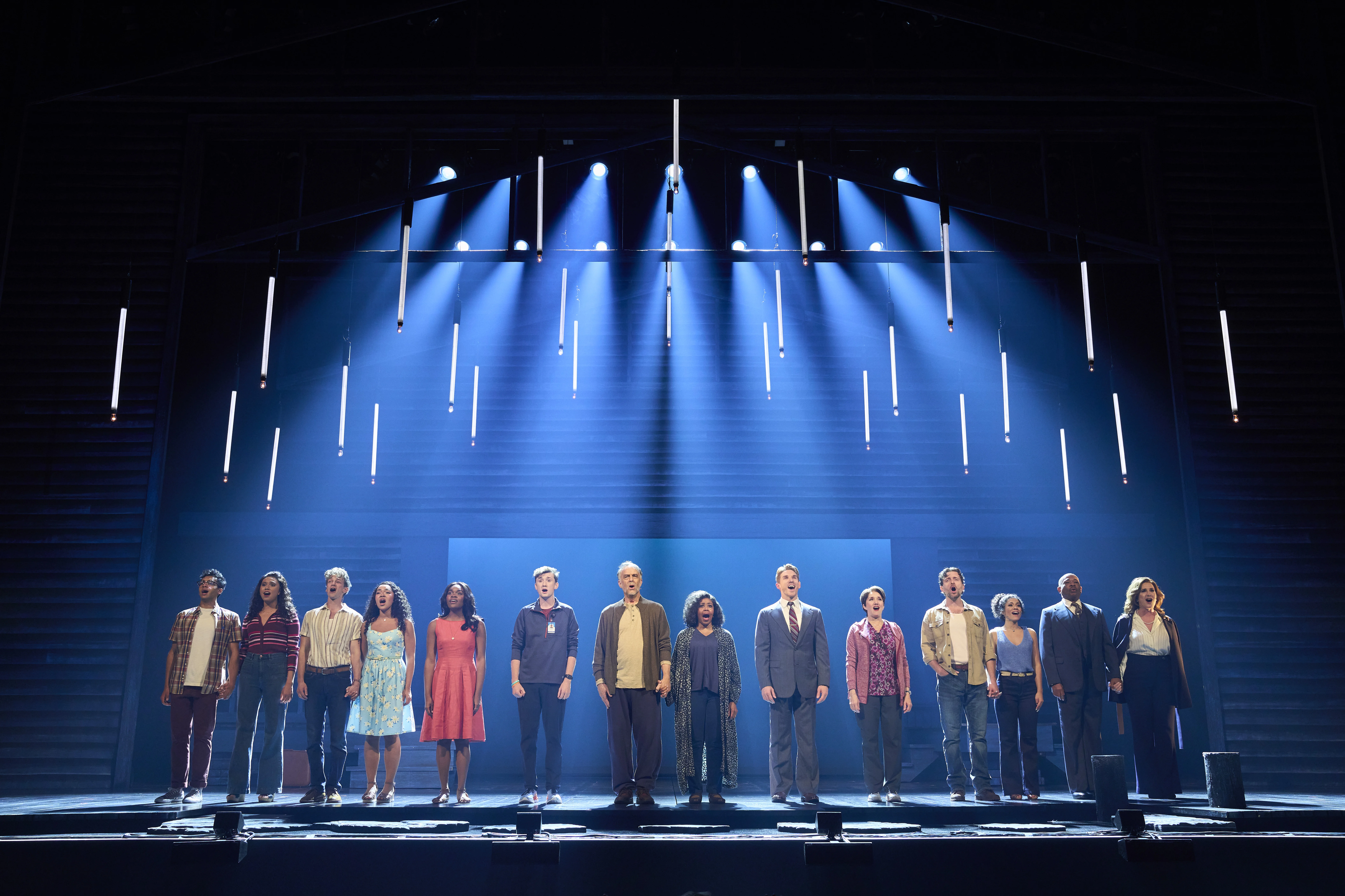 Full cast standing in a line on stage under dramatic blue lighting and vertical hanging lights during a theatrical performance, creating a striking ensemble moment.