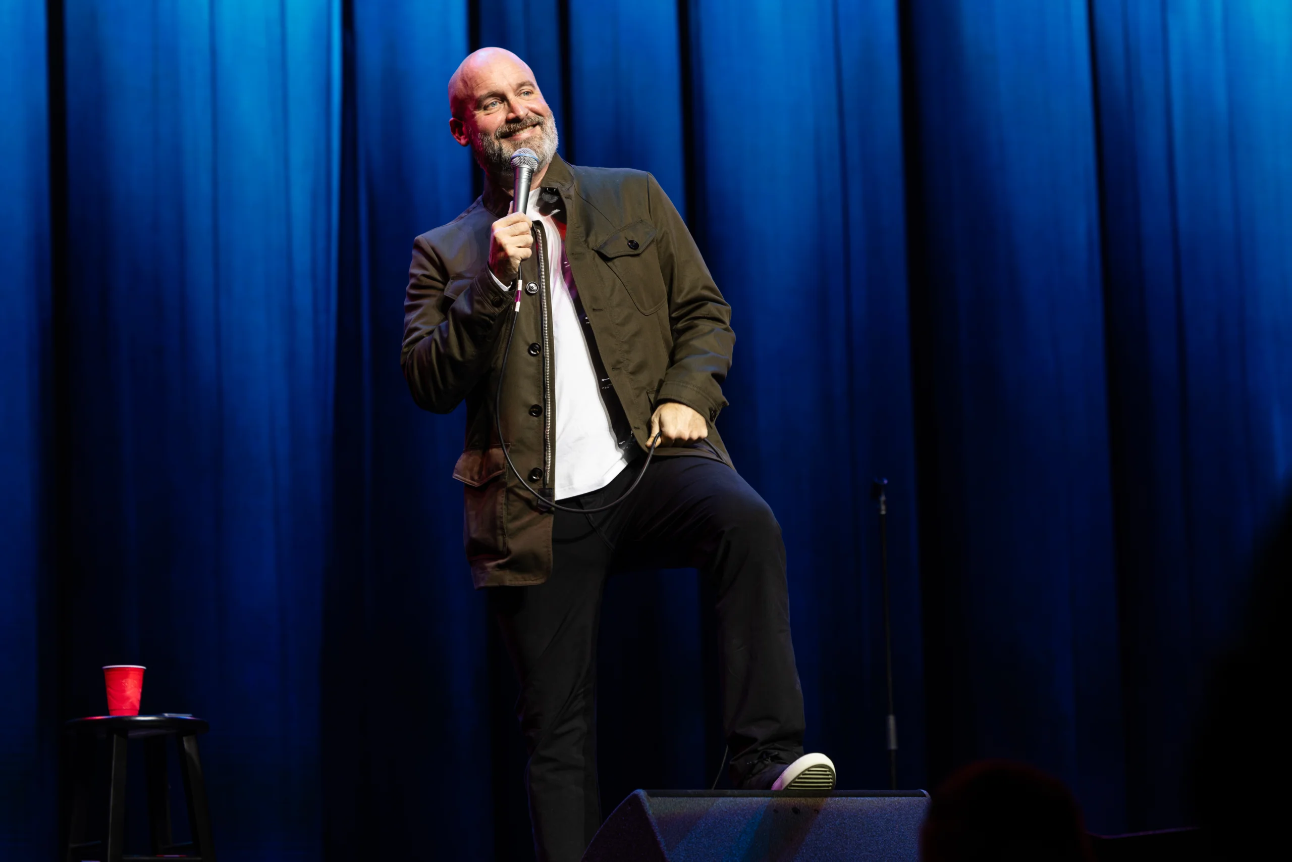 Tom Segura on stage holding a microphone, performing a stand-up comedy routine in front of blue curtains. A stool with a red cup sits to their left