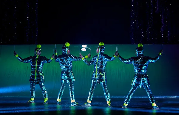 Four performers in blue metallic jumpsuits balancing lights in neon green.