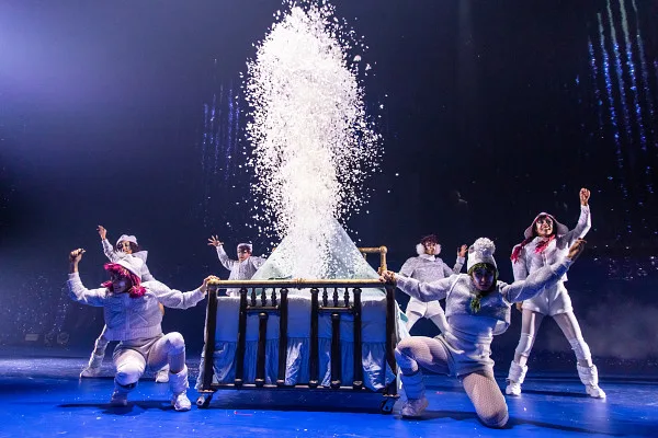 Company in white standing around a bed frame erupting with a pile of snow on it.