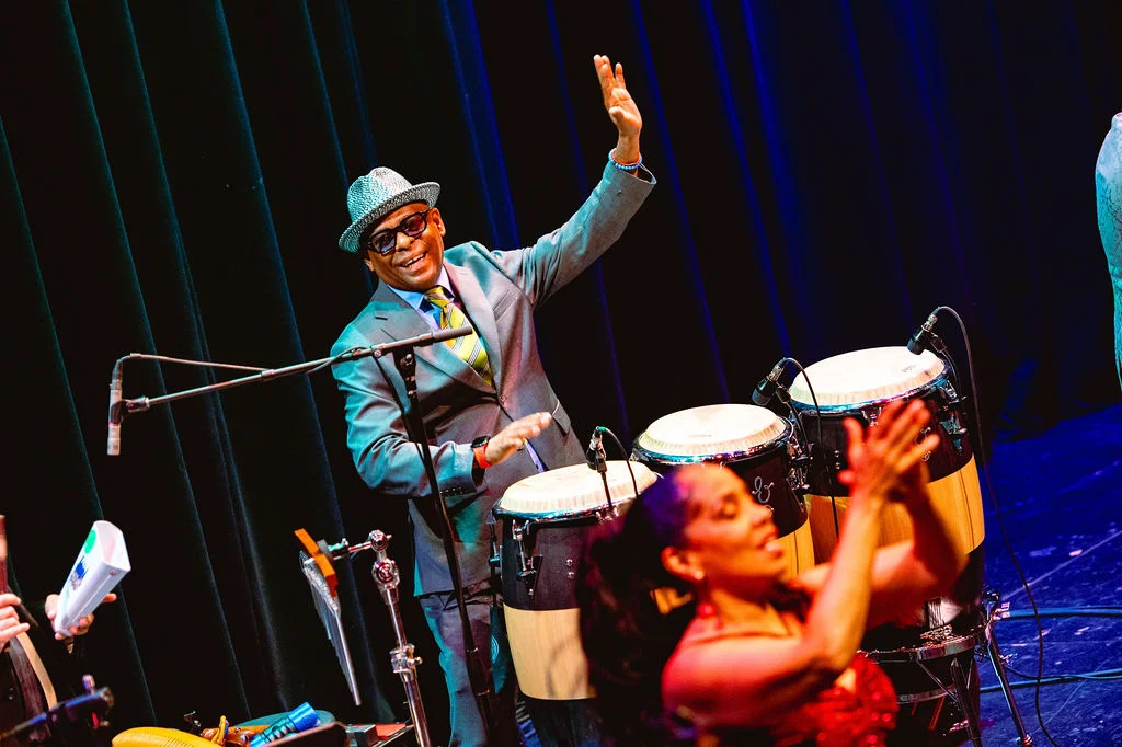 Live performance with a conga player in a suit and hat, and another person dancing or clapping in the foreground.