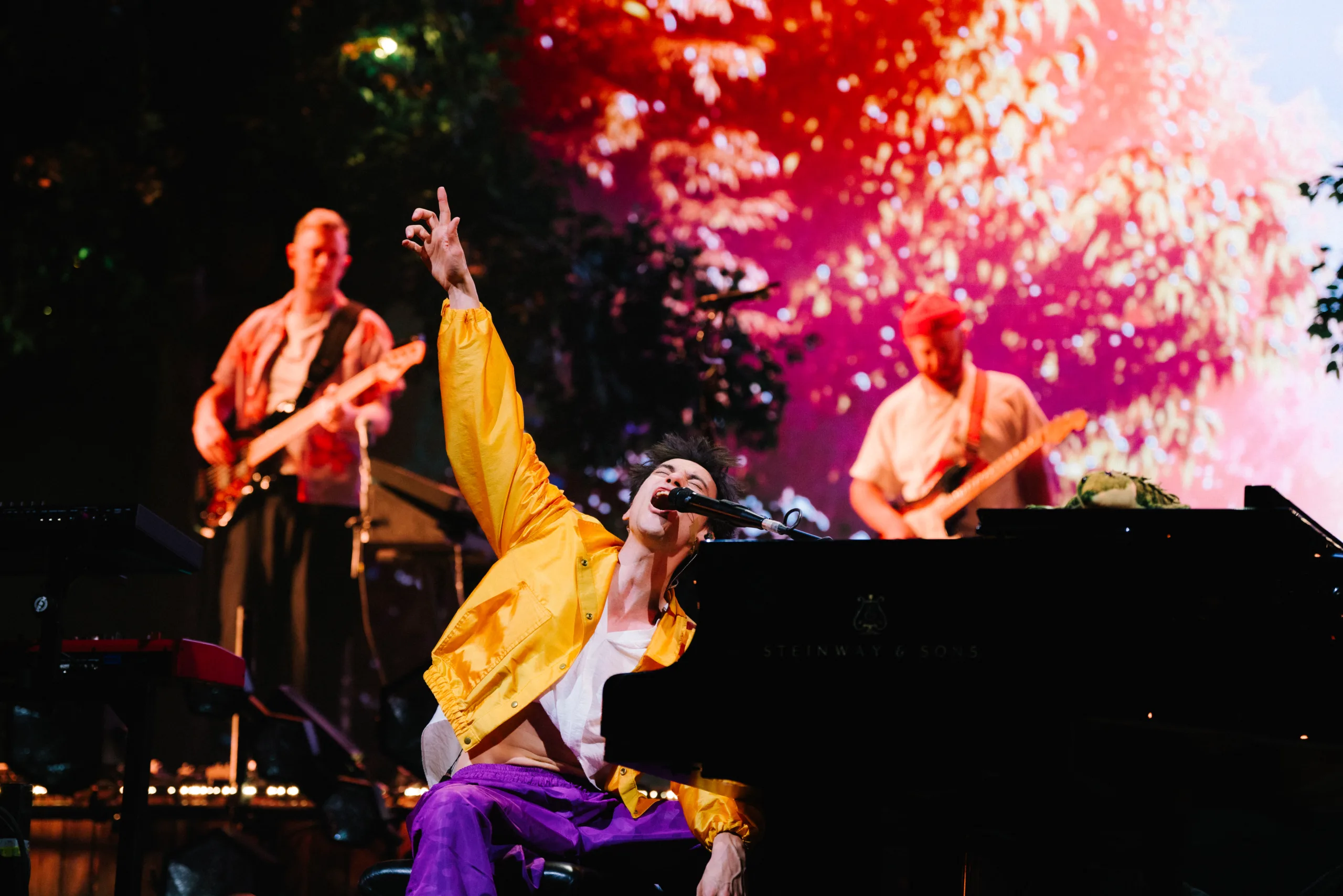 Jacob Collier on stage in a yellow jacket and purple pants passionately singing into a microphone at a grand piano with other performs in the background playin guitar and bass.