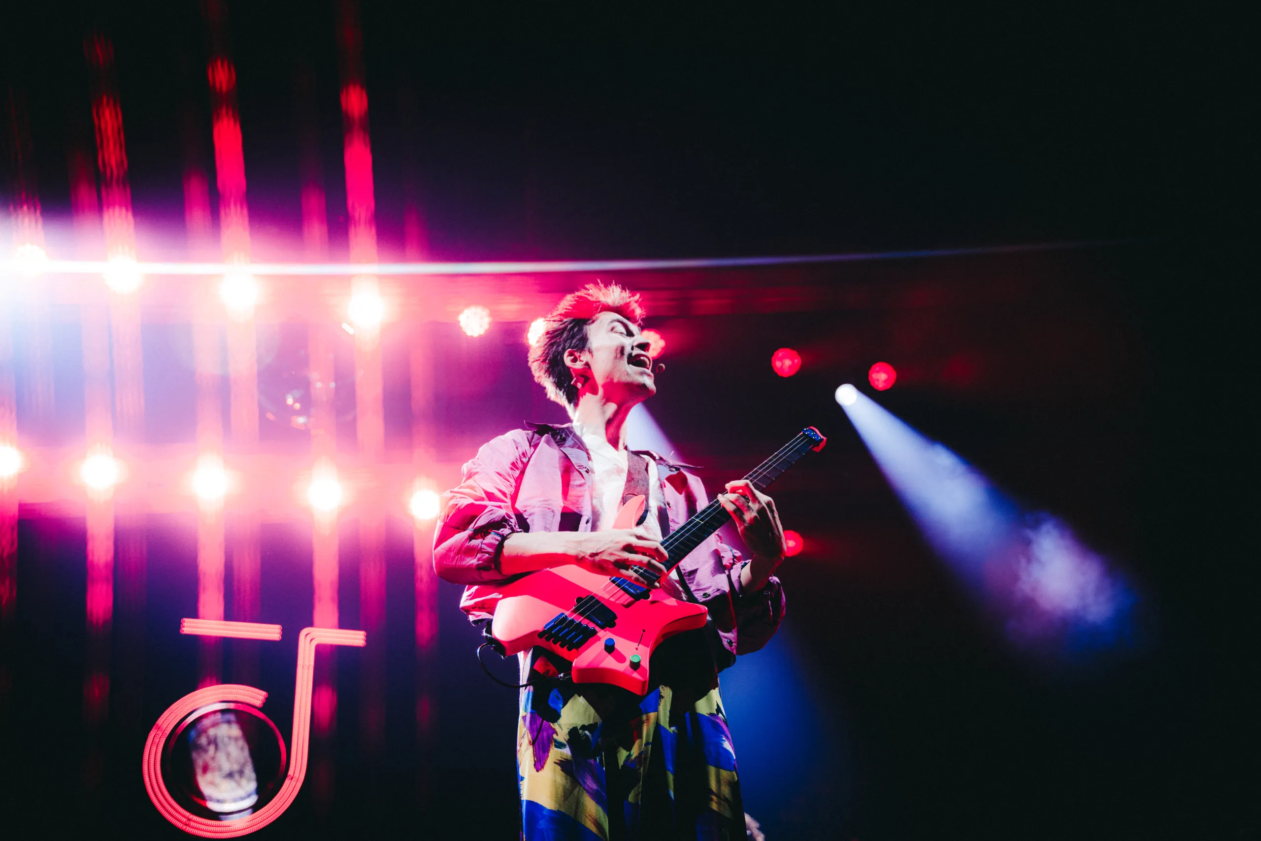 Jacob Collier on stage in a purple jacket and colorful pants holding a bright pink electric guitar with bright lights and spotlights behind him.