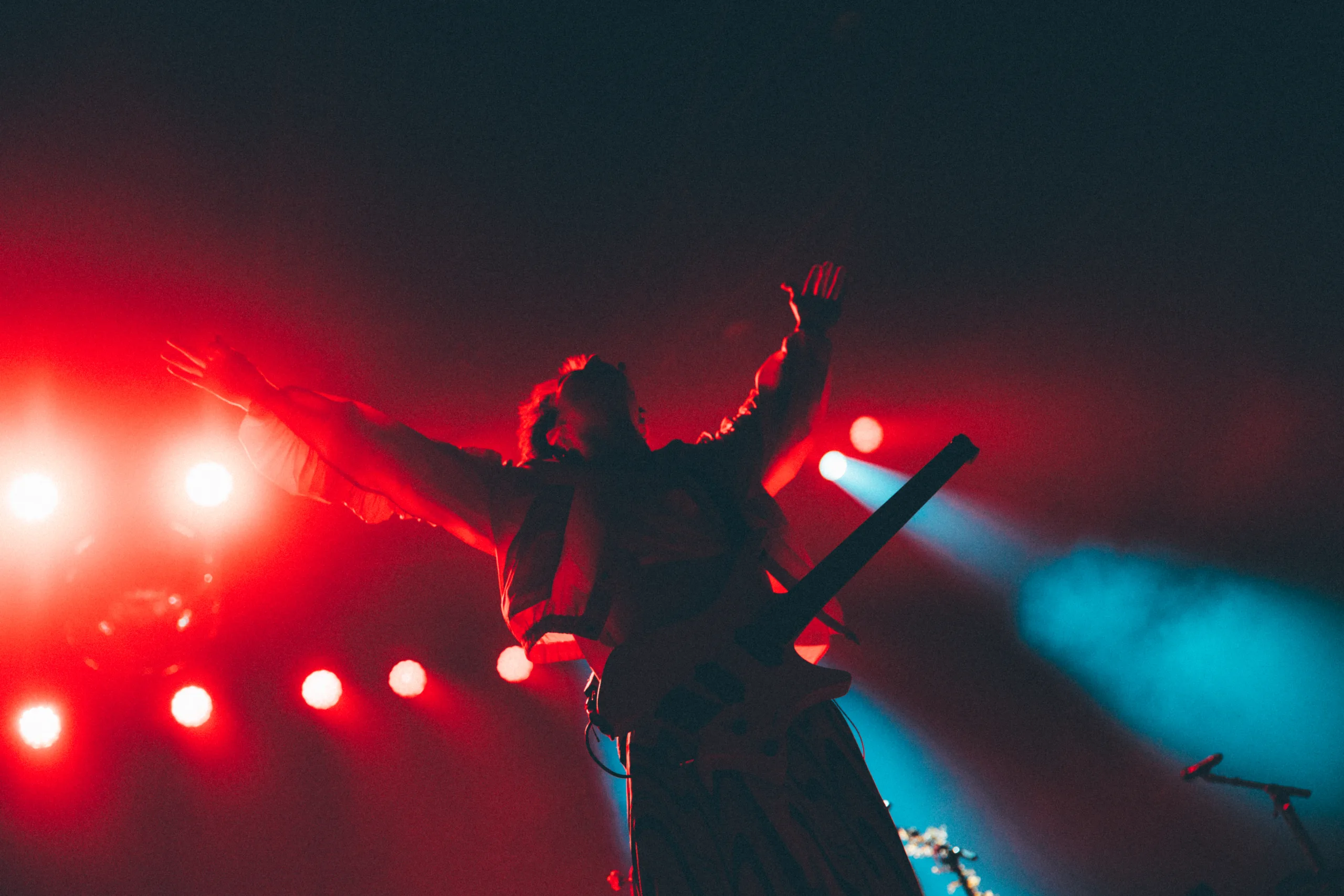 Jacob Collier silhouetted on stage with his arms outstretched ahead of hazy red lights and an electric guitar slung over his shoulders.