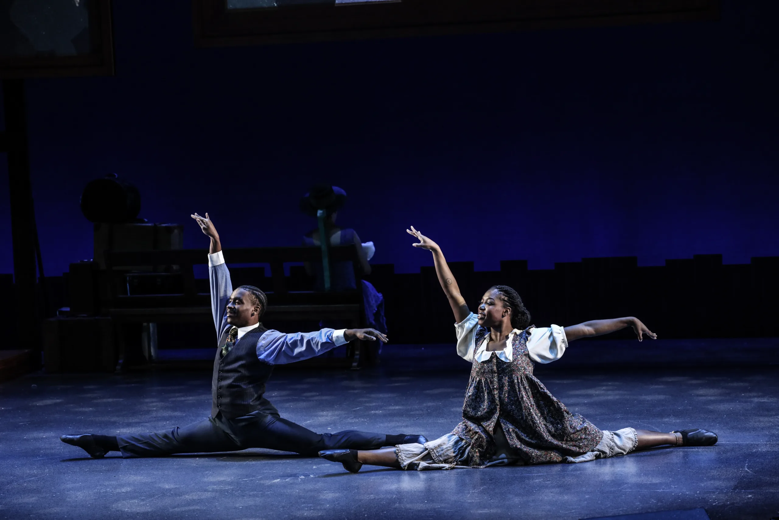 A male and female dancer in the splits on stage with arms extended.