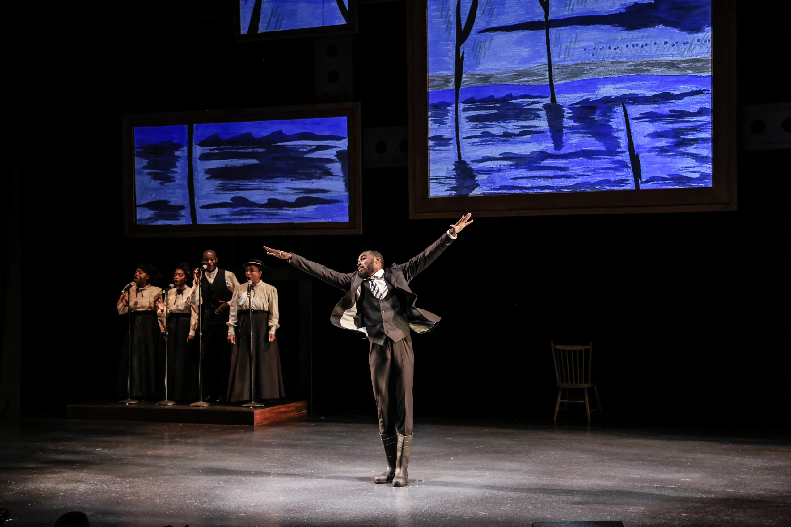 Man center stage wearing a three-piece suit with his arms flying out by his sides and four singers at microphones in the background.