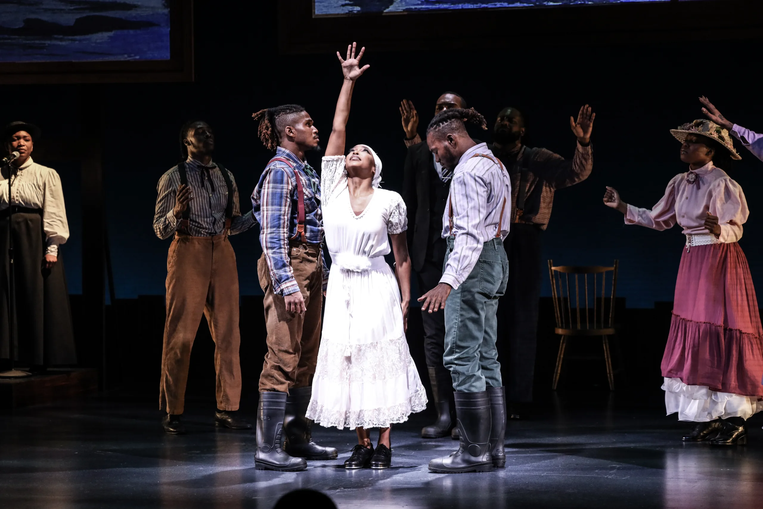 Woman center stage wearing white with her hand stretched up and performers surrounding her.