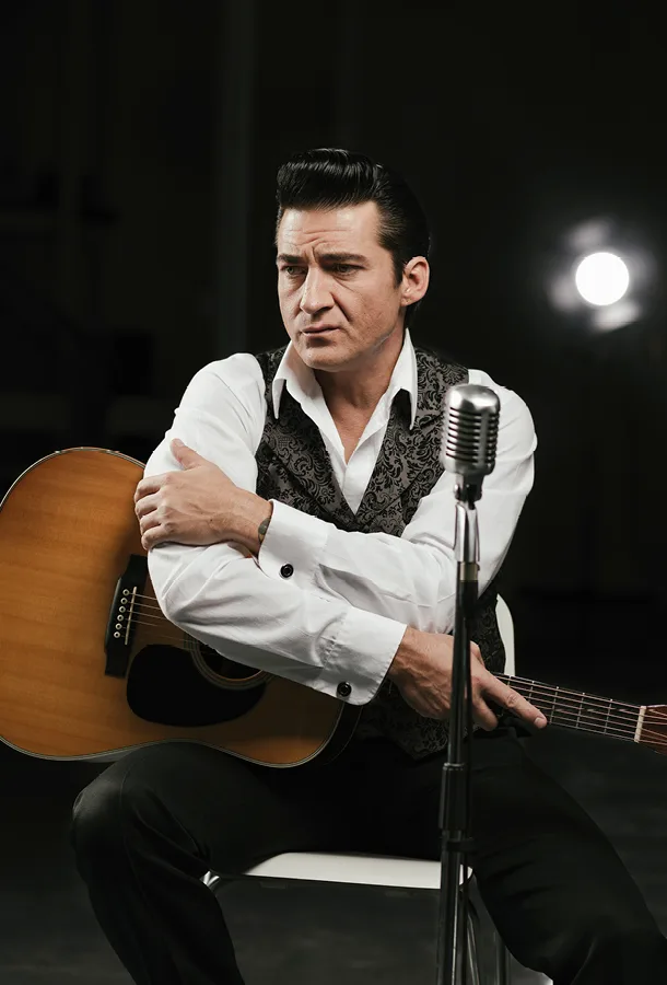Shawn Barker as Johnny Cash in front of an old fashioned microphone with his crossed arms resting on an acoustic guitar.