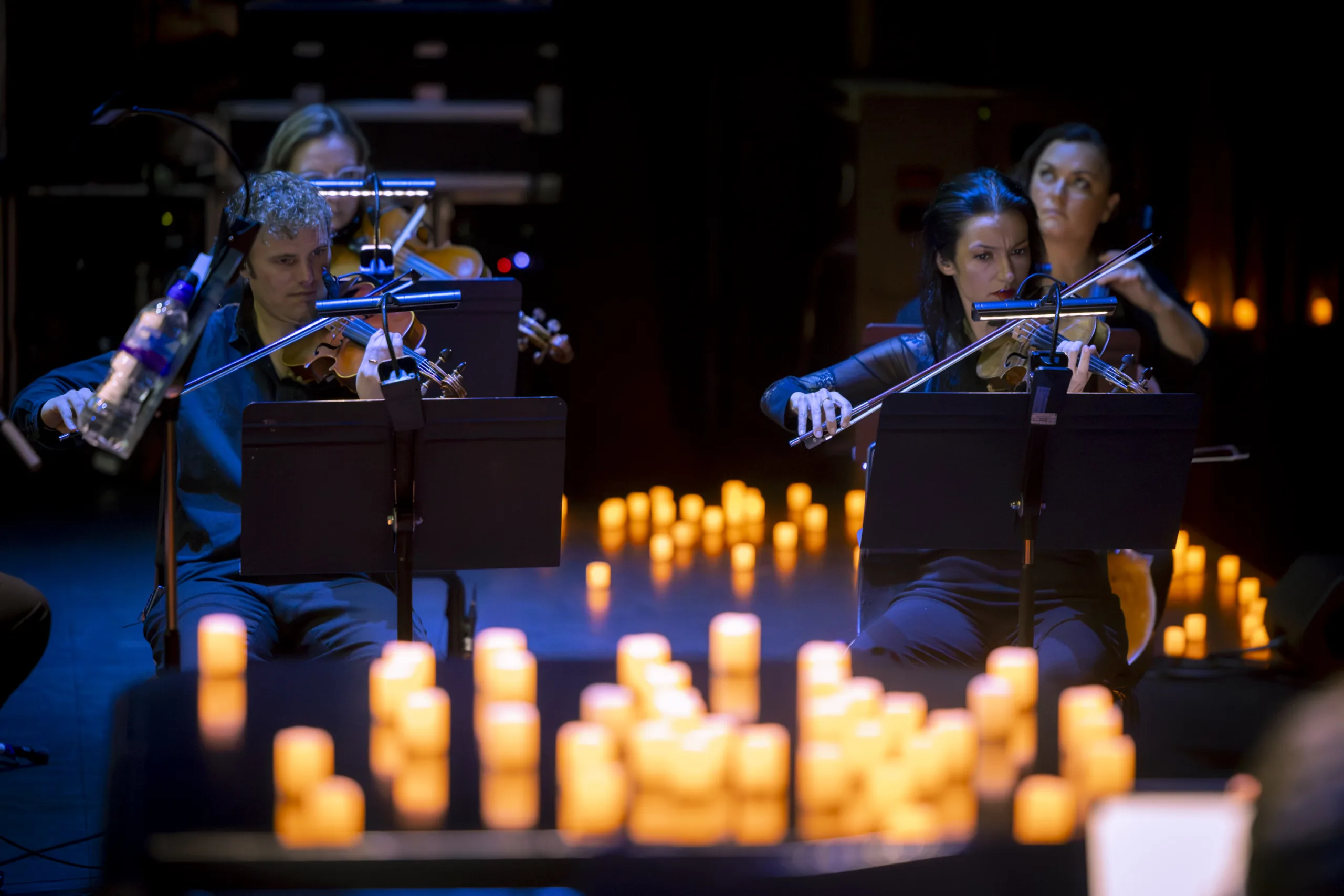 Violinists and violists playing with their music stands in front of them and lighted candles around.
