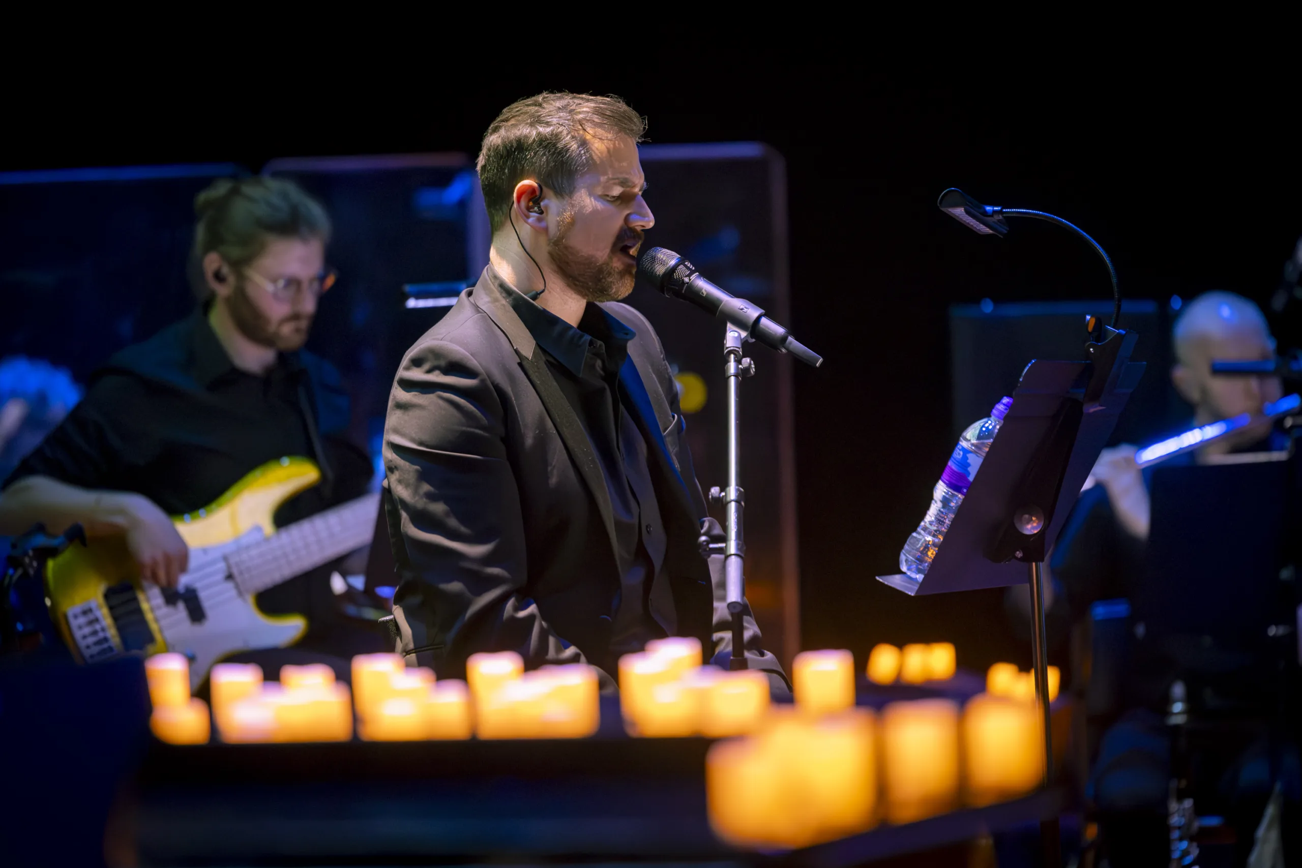 Man in a black suit singing into a microphone with his eyes closed. Candles in the foreground and a bassist and flautist in the background.