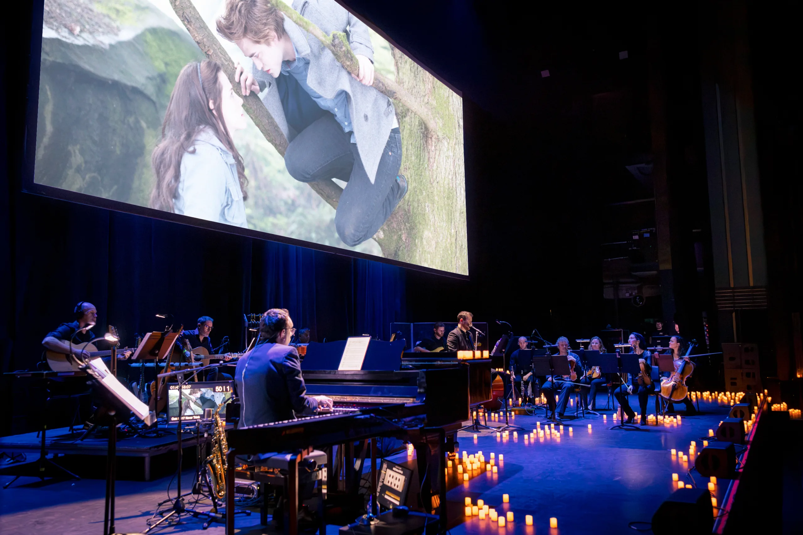 Film projection of Edward and Bella staring into each other's eyes with Edward in a tree looking down at Bella over the stage featuring the full band and a thousand candles and blue stage lights.