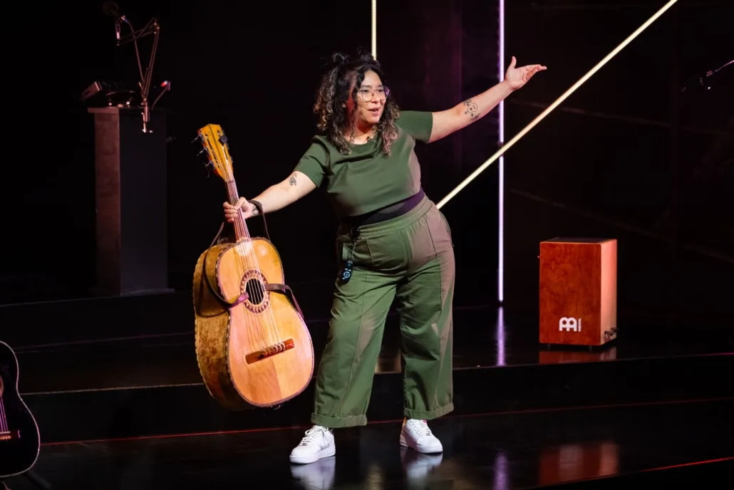 Satya Chavez, wearing a green body suit,  shows off guitar instrument on stage.